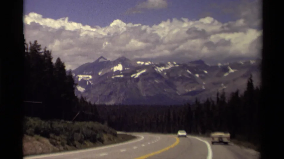 1979 Canadian mountain-drive footage with a winding highway, evergreen slopes, distant peaks, a white car, and a small trailer.