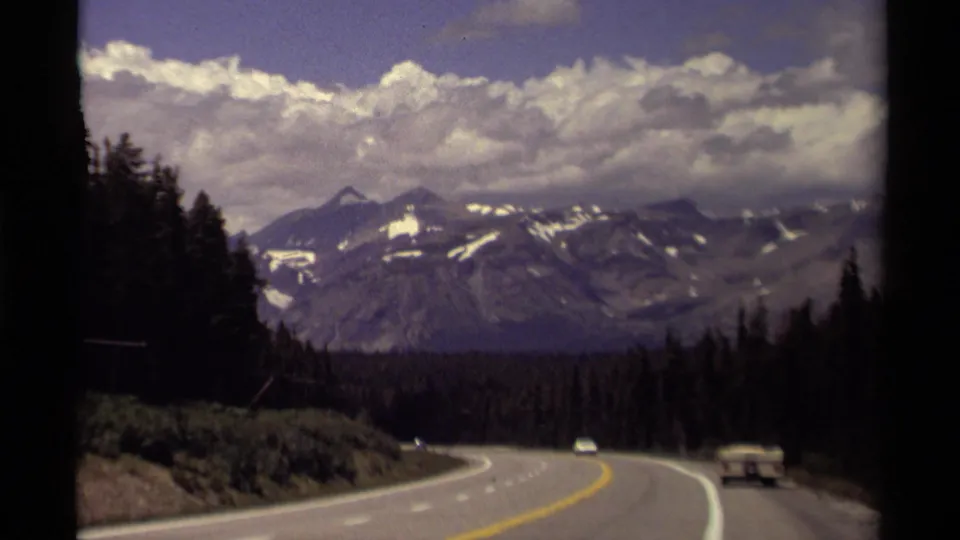 Moving-vehicle 8mm frame of a Canadian mountain road, with older cars, forested shoulders, and clouded peaks beyond.