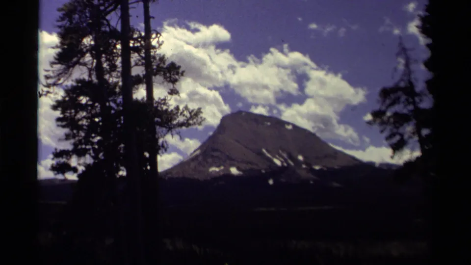 1979 Canada 8mm view of a mountain ridge with lingering snow, seen through tall dark trees below a bright cloud bank.