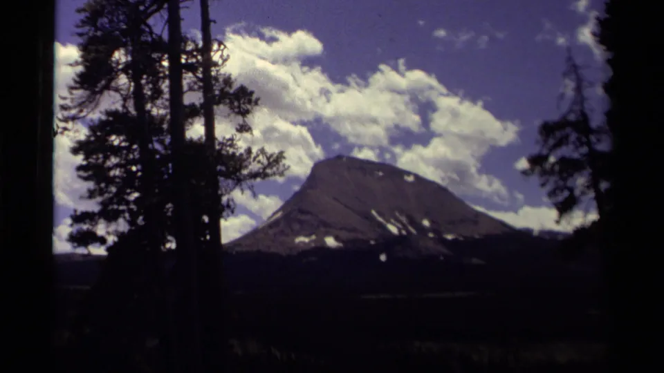 1979 Canada home-movie frame with tall trees at the edges, a dark mountain in the distance, and scattered white clouds overhead.