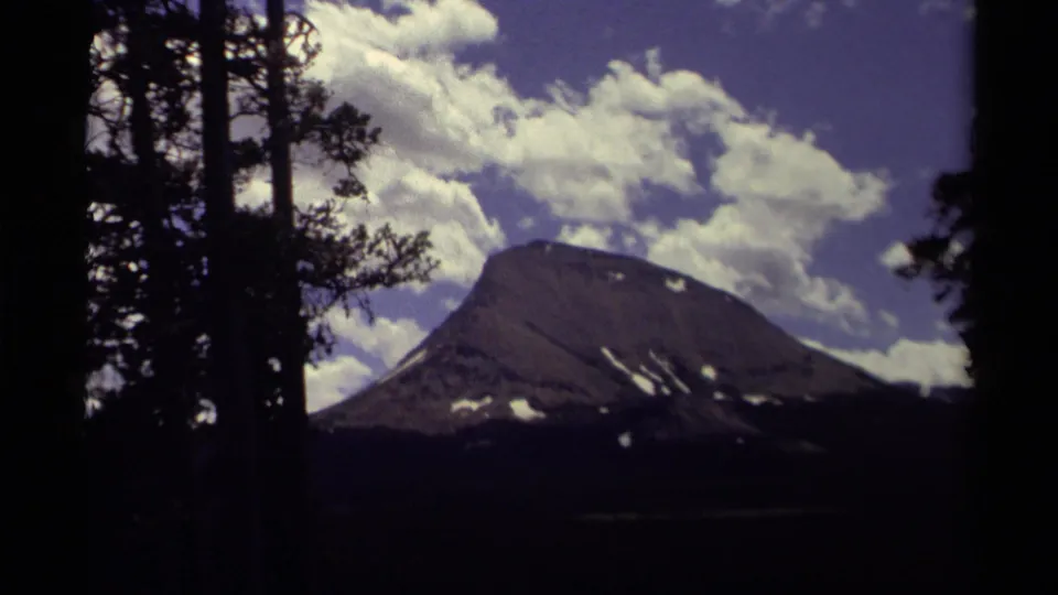 1979 Canada 8mm still of a dark mountain landscape, evergreen silhouettes, snow patches, and large white clouds against blue sky.