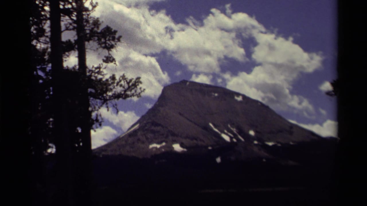 1979 Canadian 8mm frame showing a broad mountain peak with snow patches beneath a blue, cloud-filled sky and dark foreground pines.