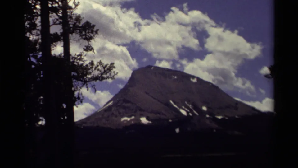 1979 Canadian 8mm frame showing a broad mountain peak with snow patches beneath a blue, cloud-filled sky and dark foreground pines.