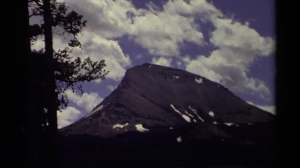 1979 Canadian 8mm frame with a snow-marked mountain peak, bright clouds, blue sky, and silhouetted trees at the edges.