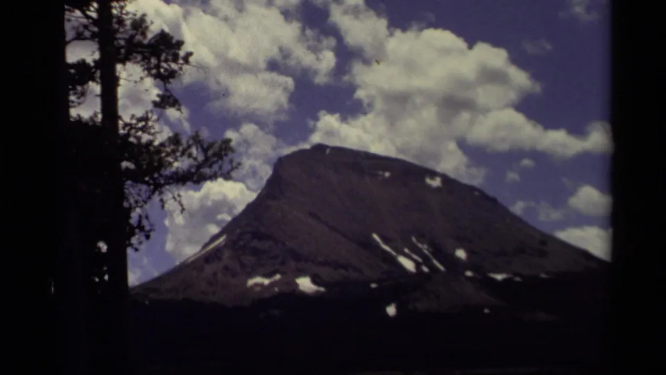 1979 Canadian 8mm frame showing the mountain slope larger in view, with snow patches, bright clouds, and dark pine silhouettes.