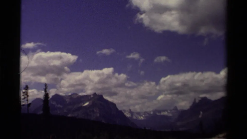 1979 Canadian 8mm frame showing distant mountain ridges, scattered clouds, blue sky, and dark tree silhouettes in the foreground.