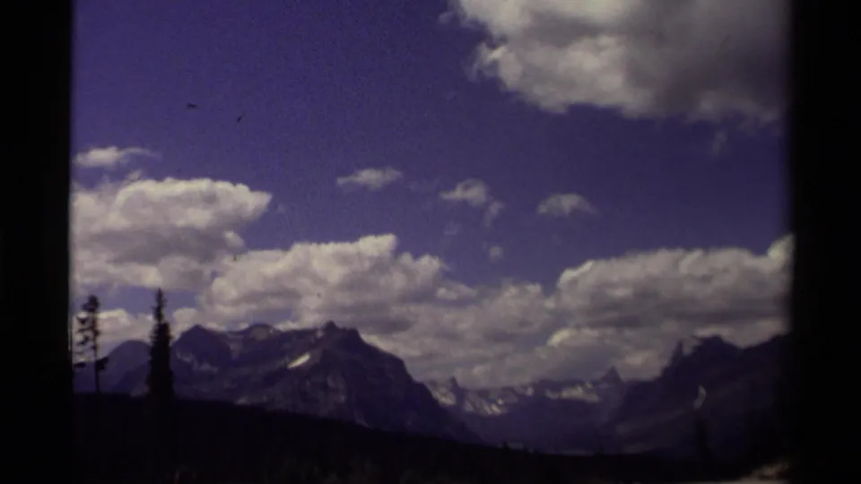 1979 Canadian 8mm frame showing rugged mountains, cloud-filled blue sky, and a dark conifer foreground within film-frame edges.