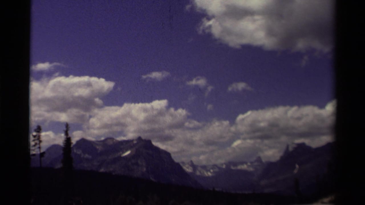 1979 Canadian home-movie frame of a mountain range beneath bright clouds, bordered by dark trees and 8mm frame edges.