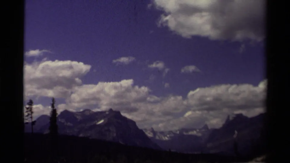 1979 Canadian home-movie frame of a mountain range beneath bright clouds, bordered by dark trees and 8mm frame edges.