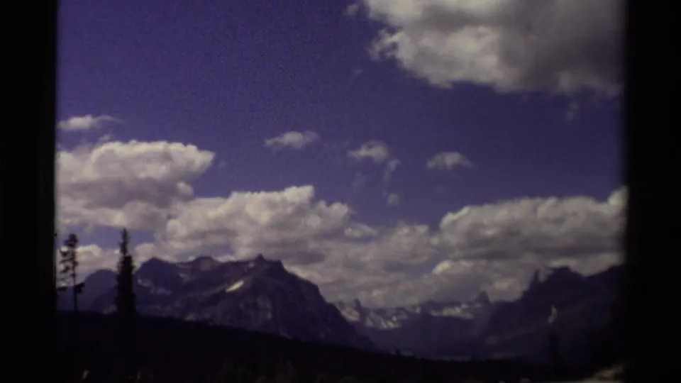 1979 Canadian 8mm frame showing distant peaks, a clouded blue sky, and dark conifers across the lower foreground.