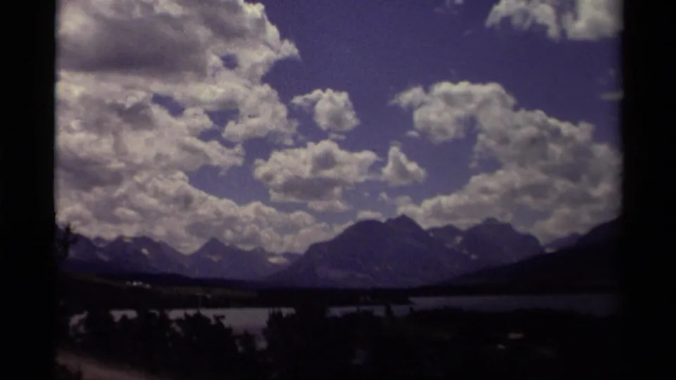 1979 Canadian 8mm frame with a wide clouded sky, mountain silhouettes, lake water, and dark forest shapes along the lower edge.