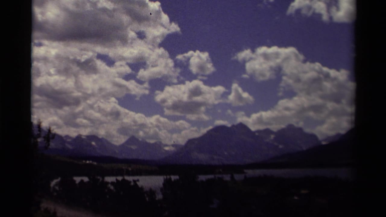 Lake and mountain range under large clouds, framed by dark film edges and foreground trees.