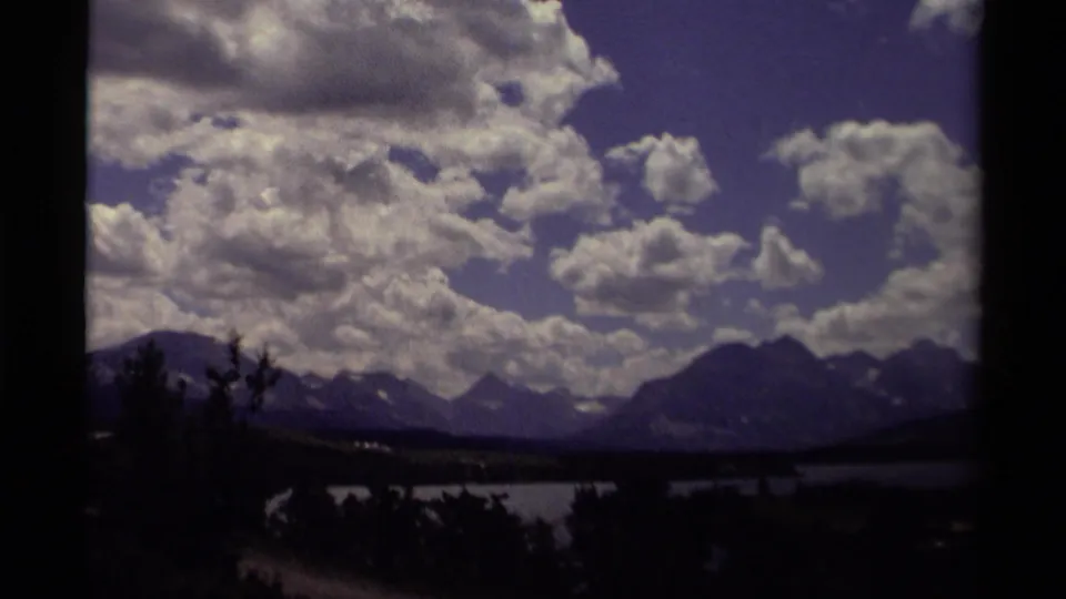 1979 Canadian 8mm landscape with a low lake, dark foreground trees, distant peaks, and a dramatic cloud bank.