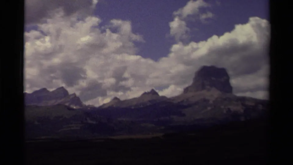 1979 Canadian 8mm frame showing a prominent block-shaped peak, ridgelines, forested slopes, and bright clouds.