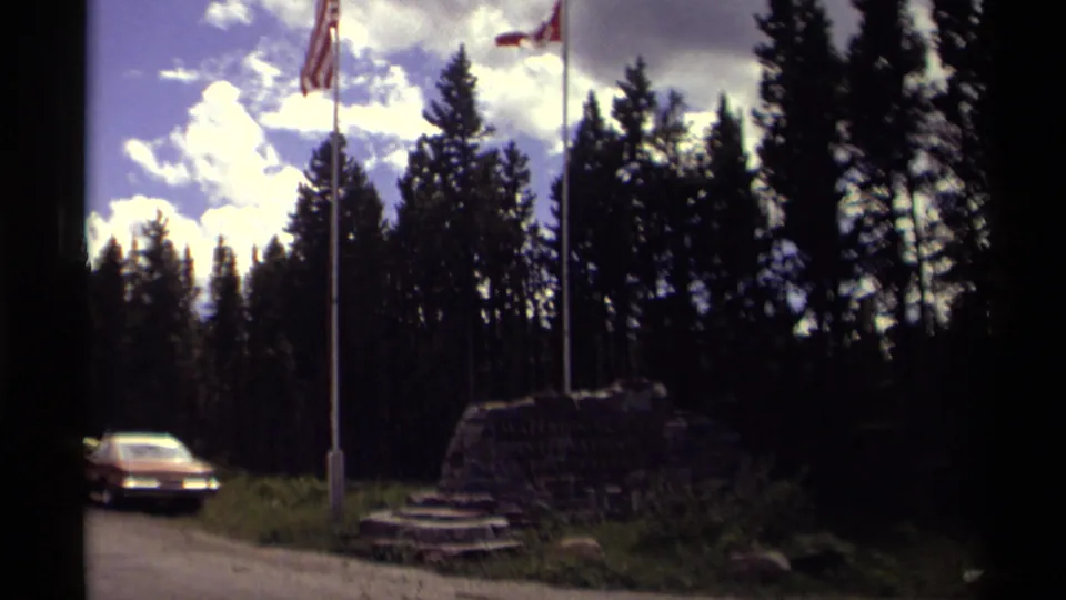 1979 Canadian roadside view with American and Canadian flags above a rock monument, evergreen trees, and a passing orange car.
