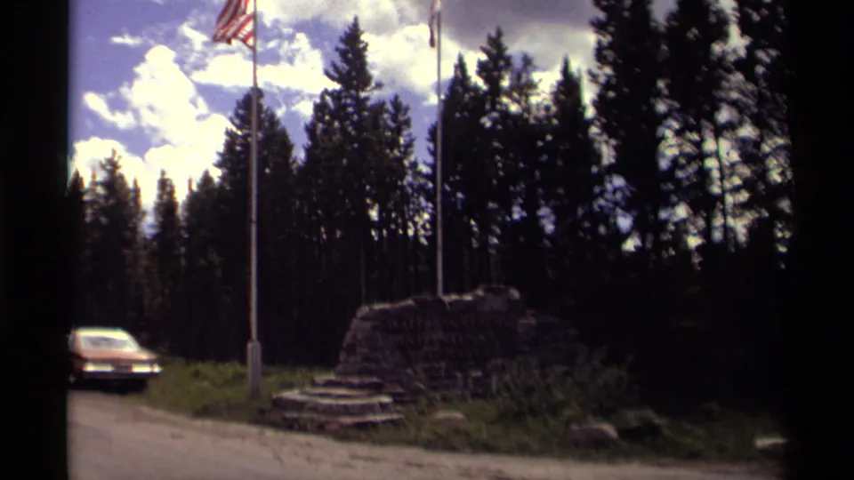 1979 Canada travel frame showing two flags over a rock monument, tall trees, cloudy sky, and an orange car on the roadside.