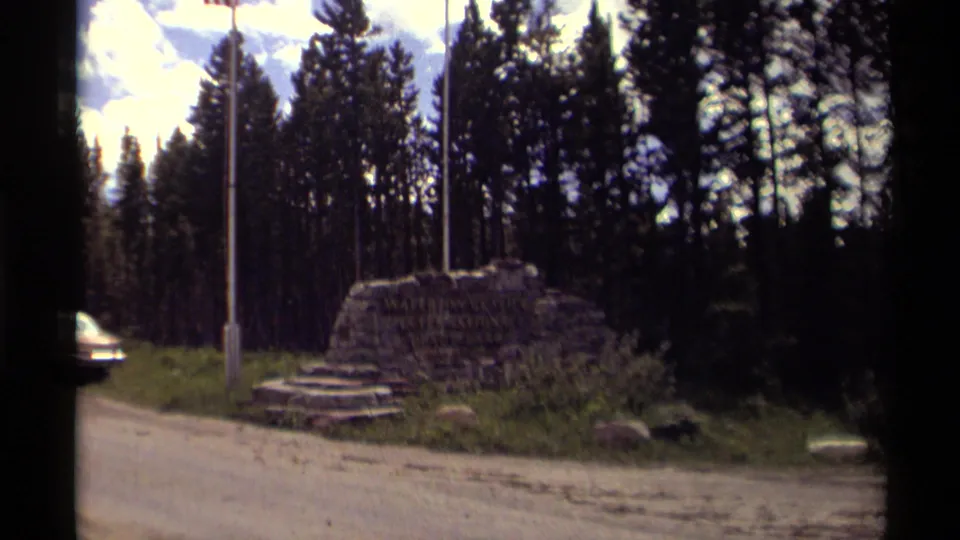 1979 Canadian roadside monument framed against evergreen trees, with flagpoles, stone steps, and a partly visible car at the road edge.