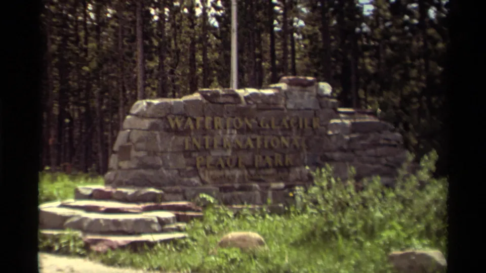 Closer 1979 home-movie view of the Waterton Glacier International Peace Park stone sign with roadside vegetation and forest behind it.