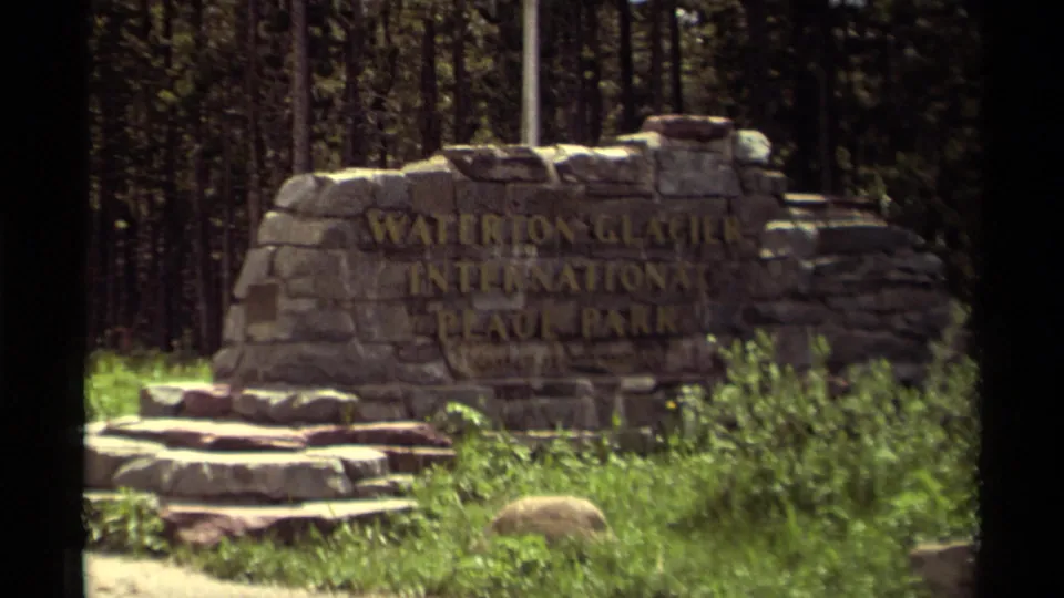 1979 8mm frame showing the Waterton Glacier International Peace Park sign more centrally, with stone steps, grass, and forest visible.