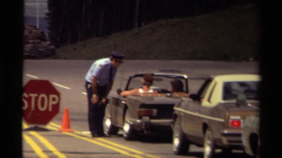 1979 Canada roadside checkpoint with a uniformed police officer beside a convertible, a red stop sign, cones, and another car waiting.