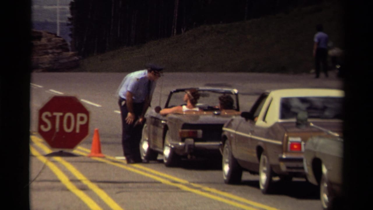 1979 Canada 8mm frame of a uniformed attendant at a roadside stop sign with a convertible and sedan waiting near forested hills.