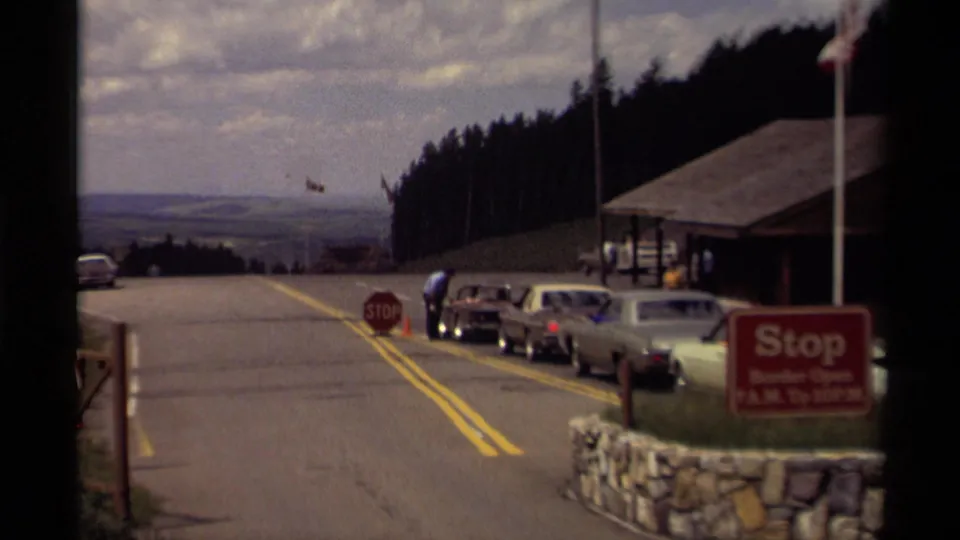 Wide 1979 Canada 8mm view of cars lined beside a rustic park building, with flags, stop signs, forest, and distant hills.