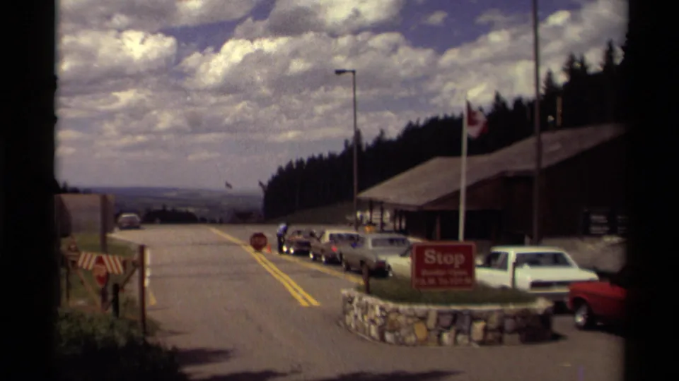 Late-1970s Canadian park entrance scene with classic cars, a stop sign, flagpoles, rustic building, forest, and broad cloudy sky.