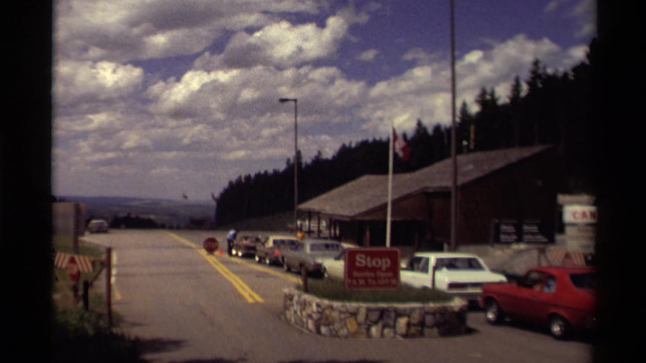 1979 Canada roadside border checkpoint with queued cars, a red stop sign, flagpole, wooden building, forest, and cloudy sky.