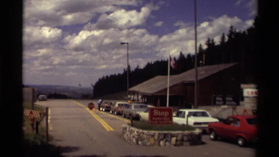 1979 Canada roadside border checkpoint with queued cars, a red stop sign, flagpole, wooden building, forest, and cloudy sky.