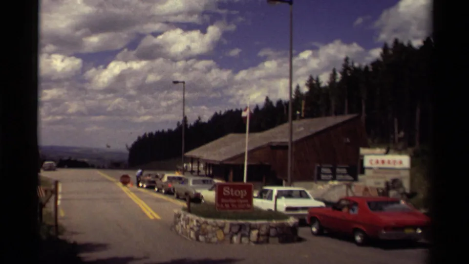 1979 Canada border approach showing classic cars near a checkpoint building, red stop sign, flags, trees, and distant landscape.