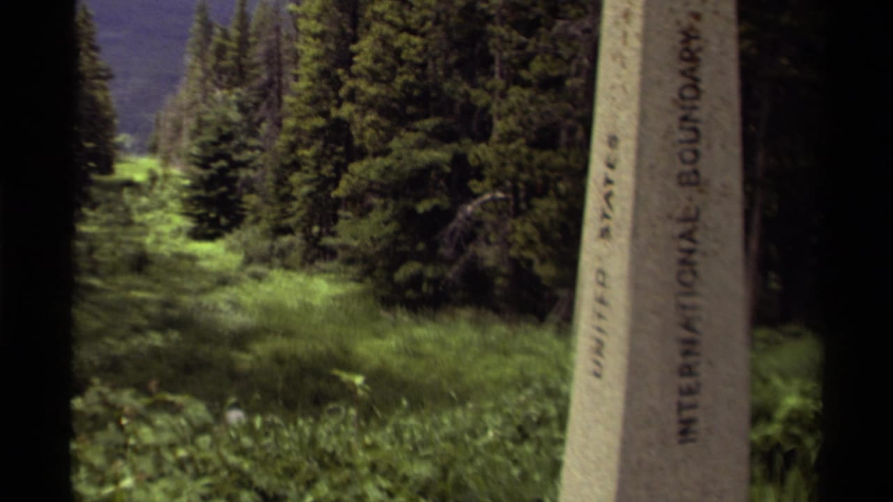 1979 Canada 8mm frame showing a stone United States-Canada boundary marker beside a grassy evergreen forest path.