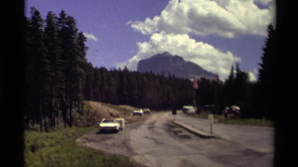 1979 Canadian roadside scene with parked cars, evergreen forest, a paved road, and a large mountain under clouds.