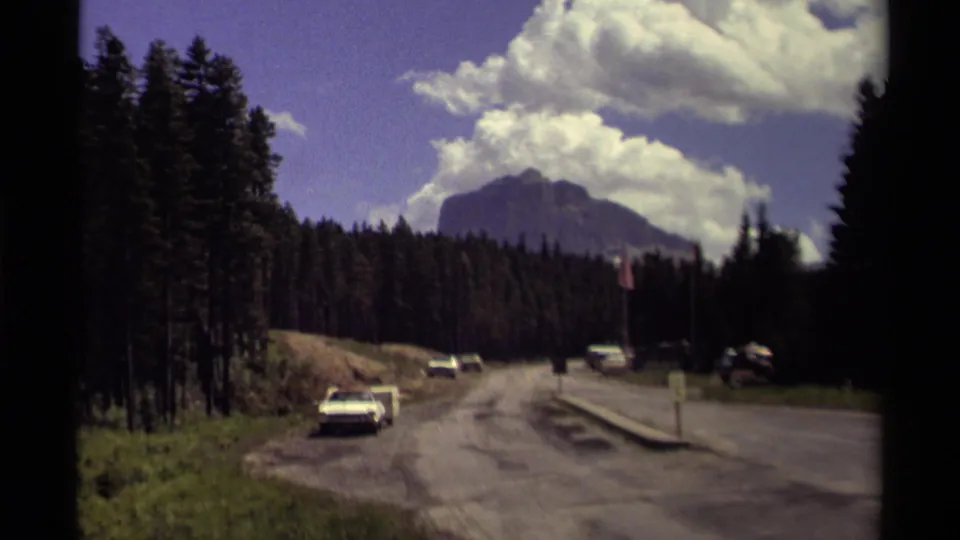 8mm home-movie frame from Canada showing parked roadside cars before evergreen forest and a mountain backdrop.
