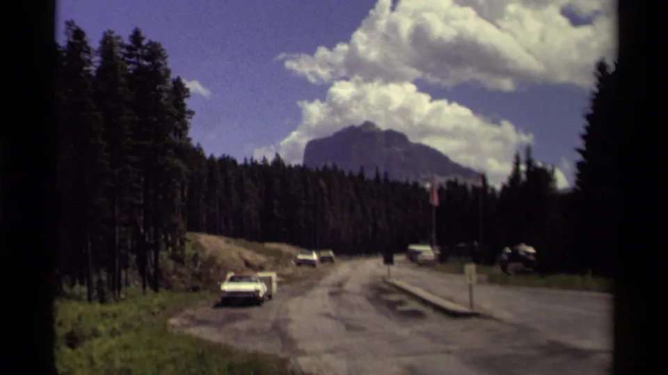 1979 8mm view of a Canadian mountain road with cars passing through forest toward a distant peak.