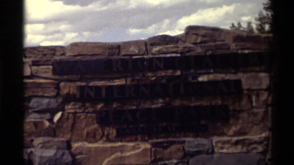 Soft-focus home-movie frame of the stone park sign, with dark lettering and cloudy sky above.
