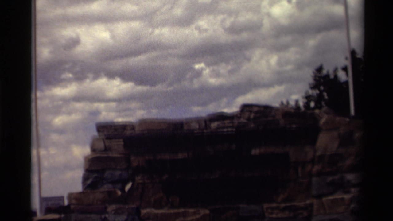 1979 Canada 8mm frame showing a shadowed cliffside rock formation below heavy clouds, with tall flagpoles near the edges.