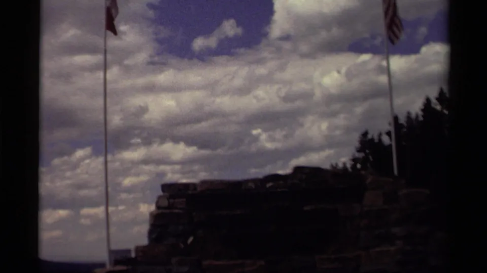 1979 Canada 8mm view of a dark cliffside rock ledge with tall flagpoles and bright clouds overhead.