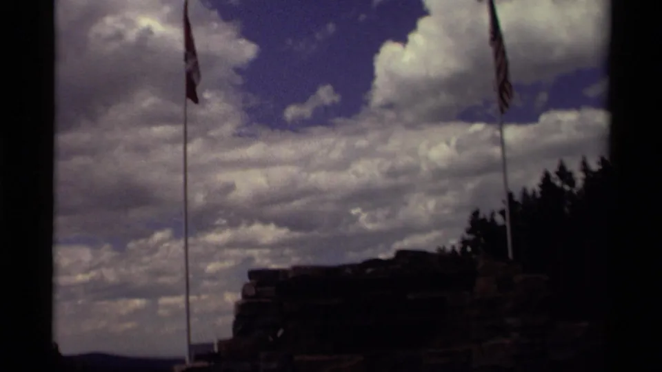 1979 Canada home-movie frame with Canadian and American flags on tall poles near a dark rock formation, trees, and dramatic clouds.