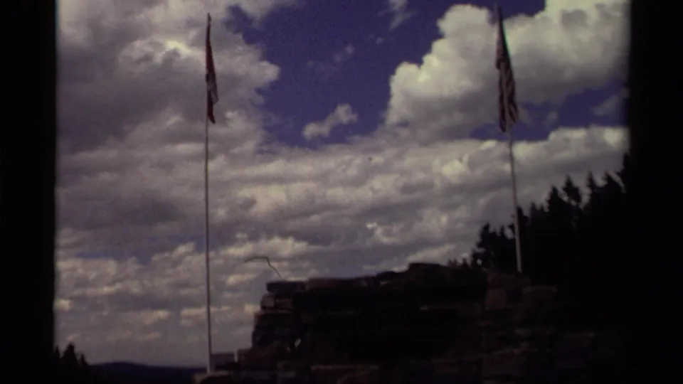 1979 Canada 8mm frame showing two flagpoles, one with an American flag, beside a shadowed rock formation and tree line.