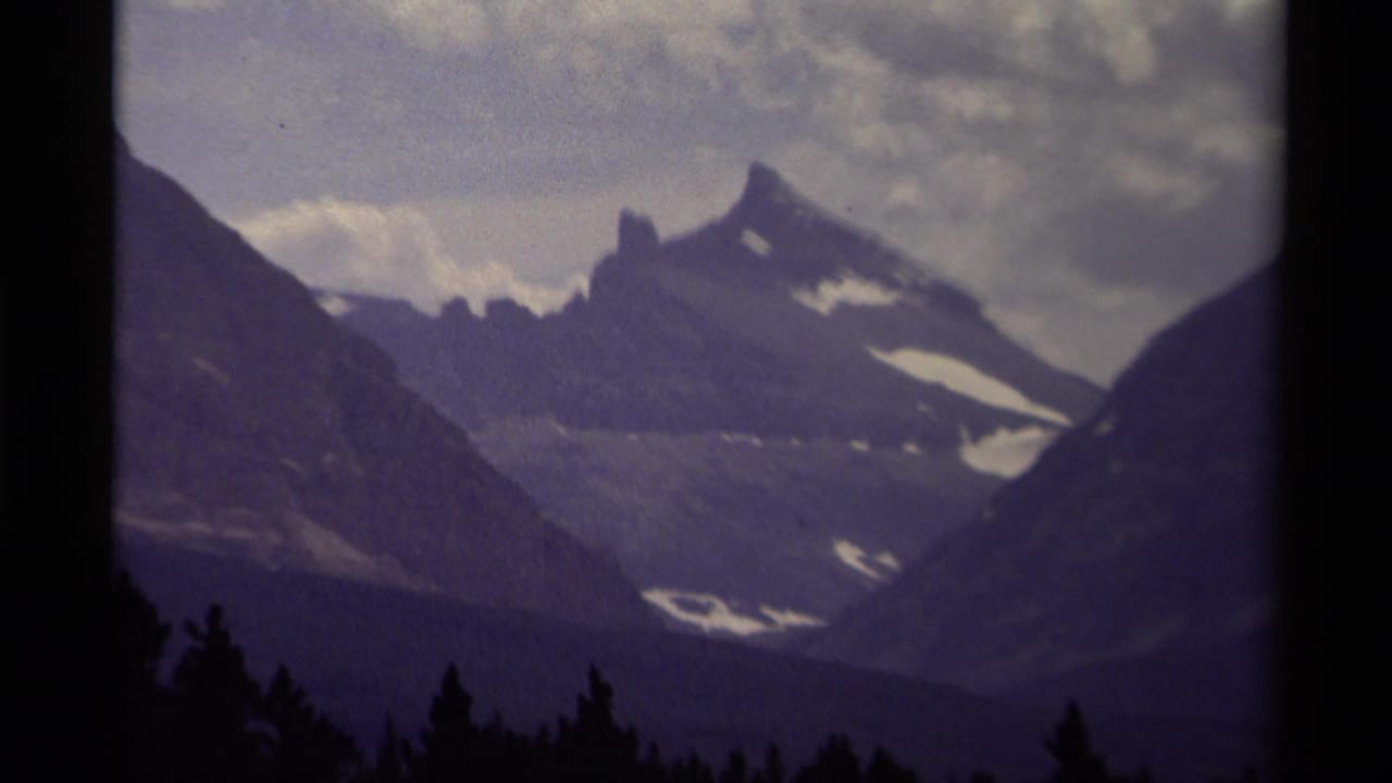 1979 Canadian 8mm frame showing a distant mountain peak with snow patches, dark forest foreground, and purple-shifted cloudy sky.