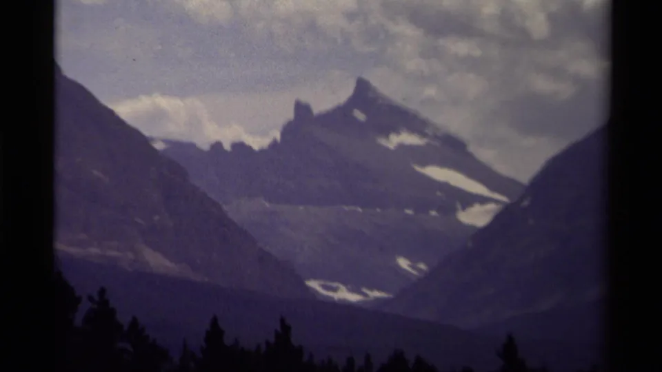 1979 Canadian 8mm frame showing a distant mountain peak with snow patches, dark forest foreground, and purple-shifted cloudy sky.