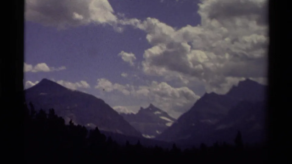 1979 Canadian 8mm frame of a wide alpine landscape, with dark conifer foreground, distant snow patches, and large clouds in a blue-purple sky.