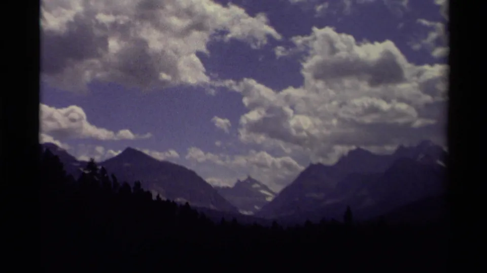 1979 Canadian 8mm frame with layered mountain slopes, a small distant snowy peak, silhouetted forest, and textured clouds overhead.