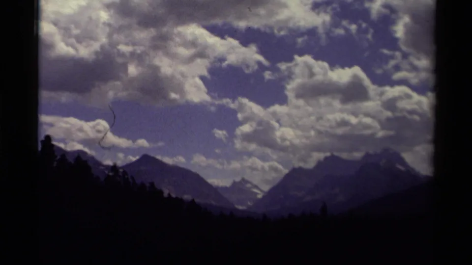 1979 Canada 8mm frame showing silhouetted trees, distant mountain peaks, snow traces, and a textured cloud-filled sky.