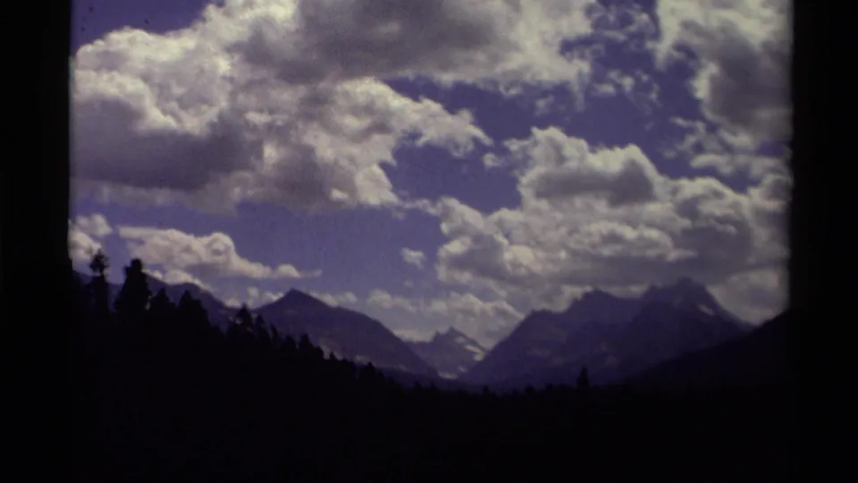 1979 Canada landscape frame with dark forest edges, layered mountain ridges, and bright cumulus clouds across a blue-purple sky.