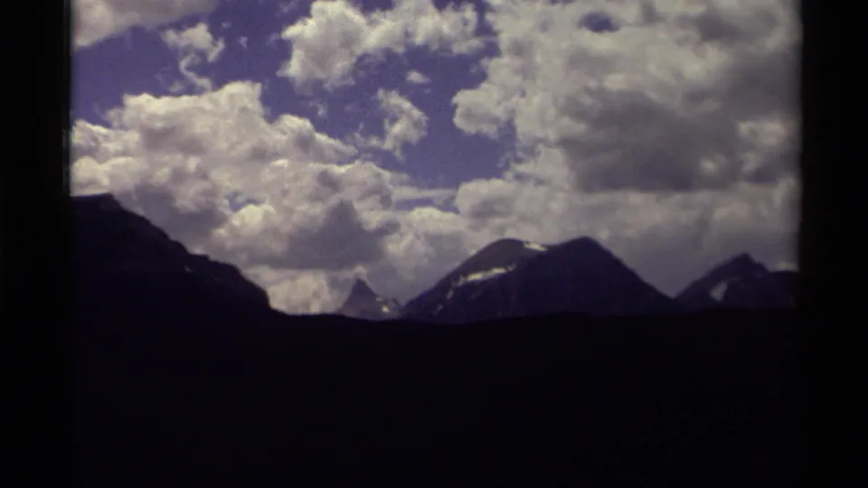 1979 Canadian 8mm frame showing distant dark mountain peaks under large white clouds and blue sky.