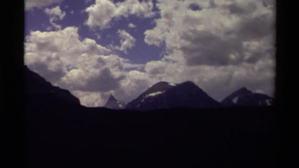 Canadian mountain range appears in dark silhouette beneath textured white clouds in faded 8mm color.