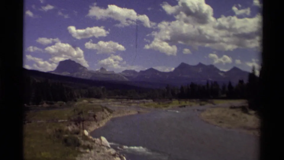 1979 Canadian home-movie frame of a river valley with dark tree line, mountain peaks, and bright summer clouds.