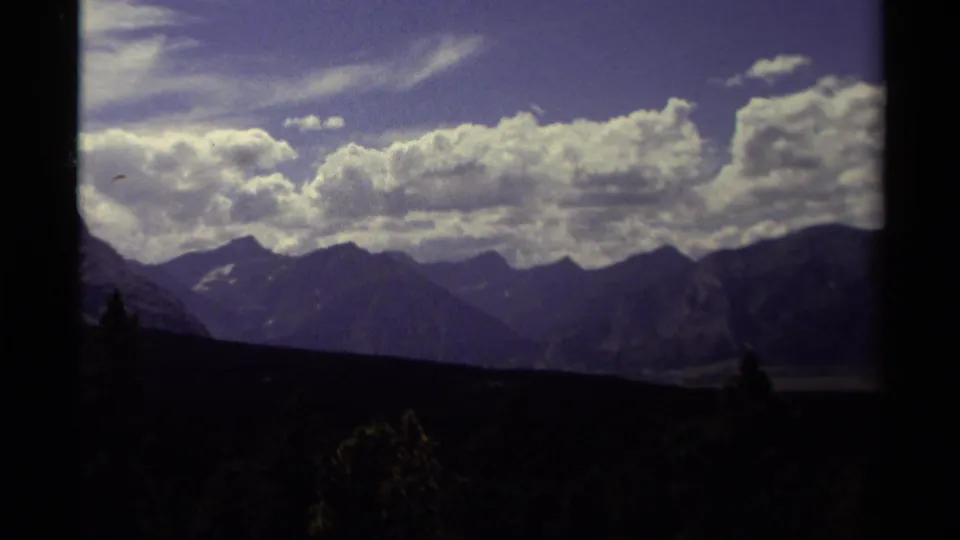 1979 Canadian home-movie frame showing ridges and a possible valley beneath bright clouds, with silhouetted trees in front.