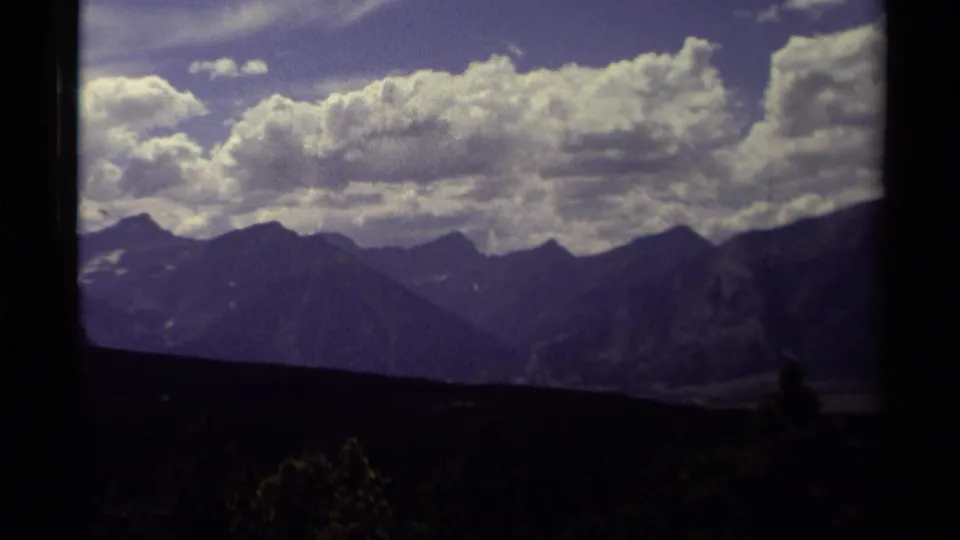 Canadian mountain range on 8mm film, with layered ridges, jagged peaks, and a bright cloud bank above the dark lower landscape.